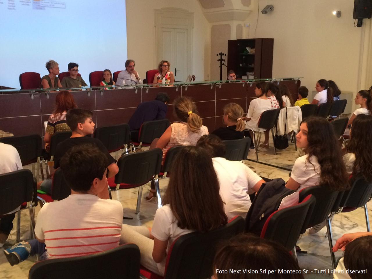 Persone sedute, donna che parla, bimba vestita da sindaco, platea